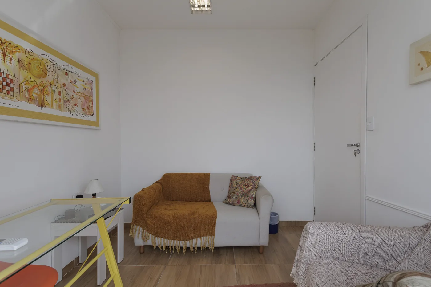 Sala Ágata, com sofá de dois lugares, manta cor de mostarda e quadro em tons de amarelo